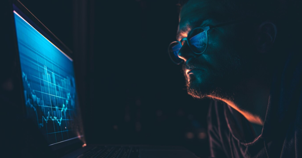 A man intently studies financial charts on a computer screen at night